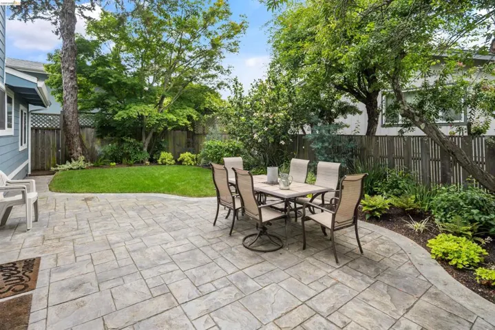 Expansive stamped concrete patio with curved edges, surrounded by mature trees, lush perimeter landscaping, and a manicured lawn