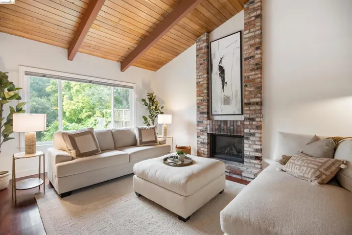 Living area featuring vaulted wood-paneled ceilings with exposed beams, a brick-surround fireplace, a large picture window, and dark wood-finish flooring