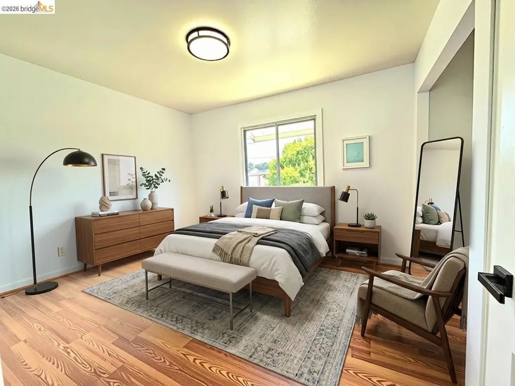 Bright room featuring warm wood-finish flooring, a large window with exterior greenery, and a contemporary flush-mount ceiling light. Virtual staging.