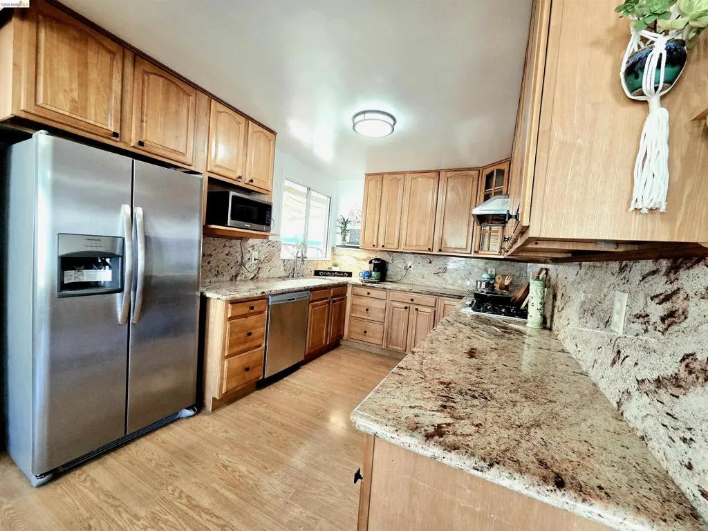 Kitchen featuring wood cabinetry, light-toned granite countertops and backsplash, stainless steel appliances, and wood-finish flooring