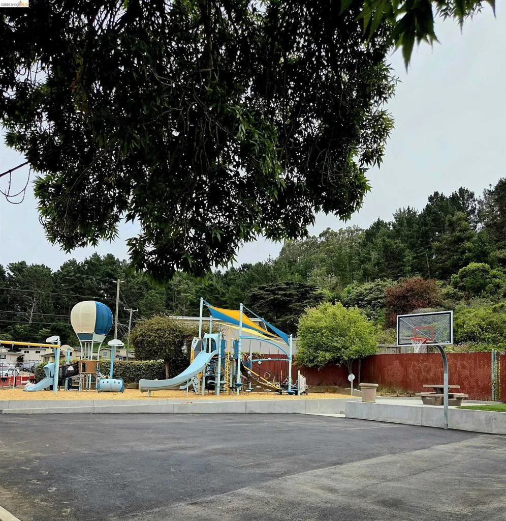 Outdoor recreational area featuring a paved basketball court, playground equipment with slides and climbing structures, and a shaded picnic table