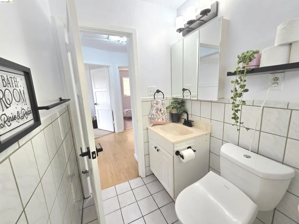 Bathroom featuring white square tile walls, white square tile flooring with contrasting grout, a white vanity with a built-in sink, a wall-mounted medicine cabinet with integrated lighting, and a white toilet