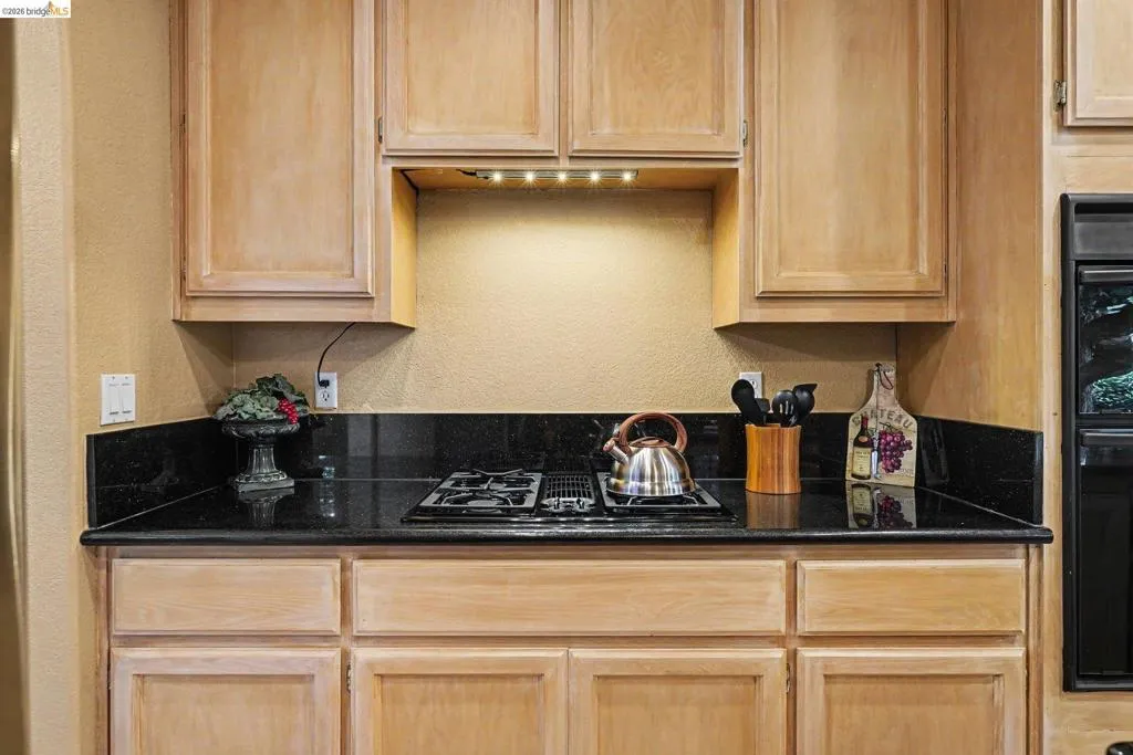 Gas cooktop with a black countertop, under-cabinet lighting, and light wood cabinetry