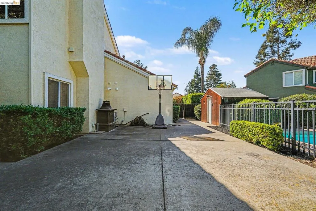 Exterior stucco finish, concrete patio, mature hedging, metal fence, and a visible pool