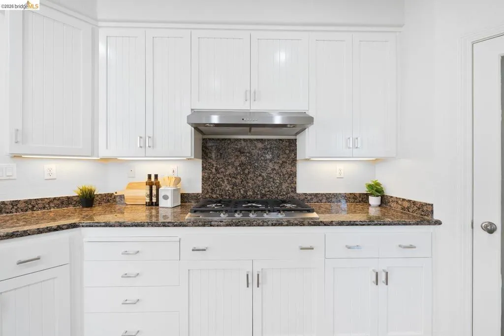 White cabinetry with silver-tone hardware, granite countertops, stainless steel range hood, built-in gas cooktop, and under-cabinet lighting