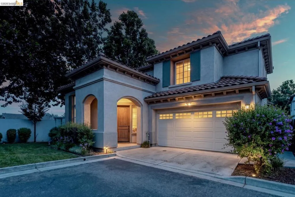 Stucco exterior featuring a tiled roof, arched entryway with a wood-finish door, attached garage with window inserts, and green landscaping