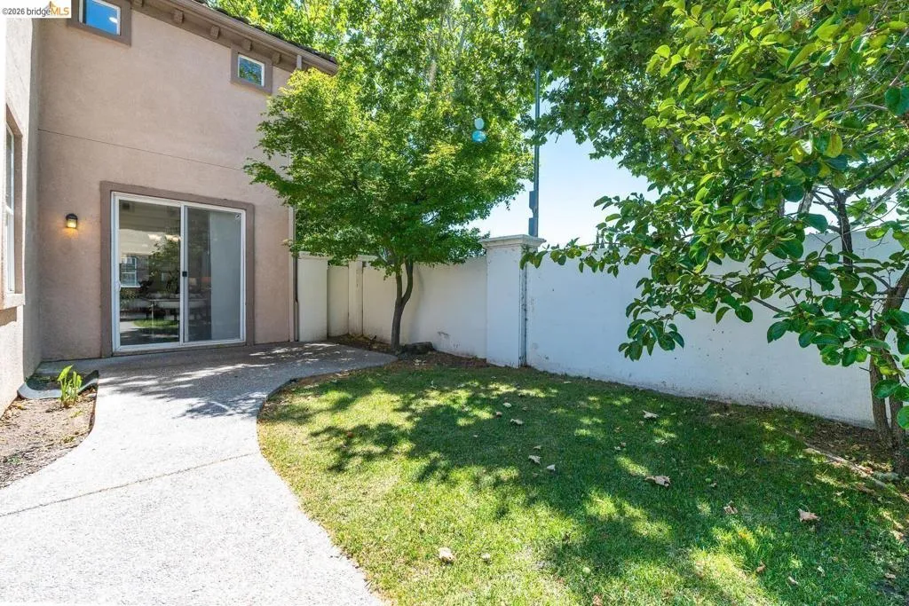 Rear exterior featuring a sliding glass door, a curved concrete pathway, a manicured lawn, and mature trees