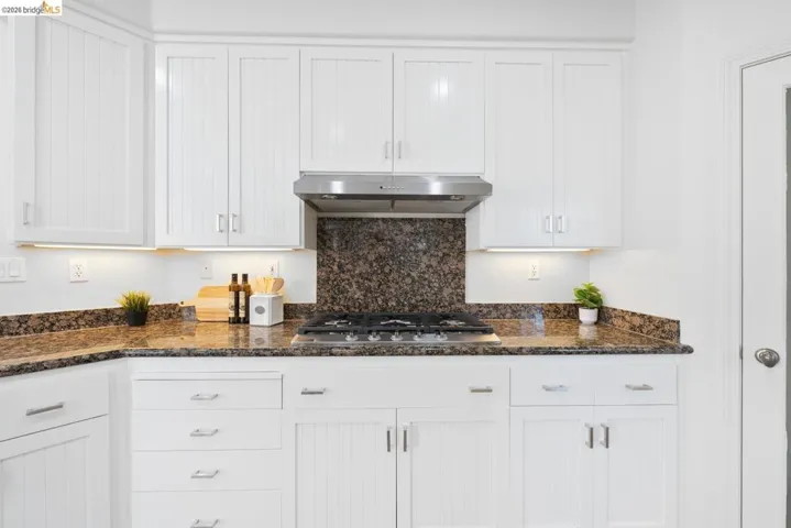 White cabinetry with silver-tone hardware, granite countertops, stainless steel range hood, built-in gas cooktop, and under-cabinet lighting