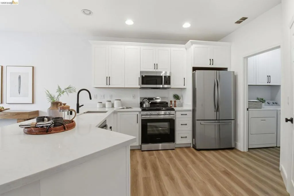 Modern kitchen featuring white shaker cabinetry, stainless steel appliances, a black gooseneck faucet, light-toned countertops, and wood-finish flooring