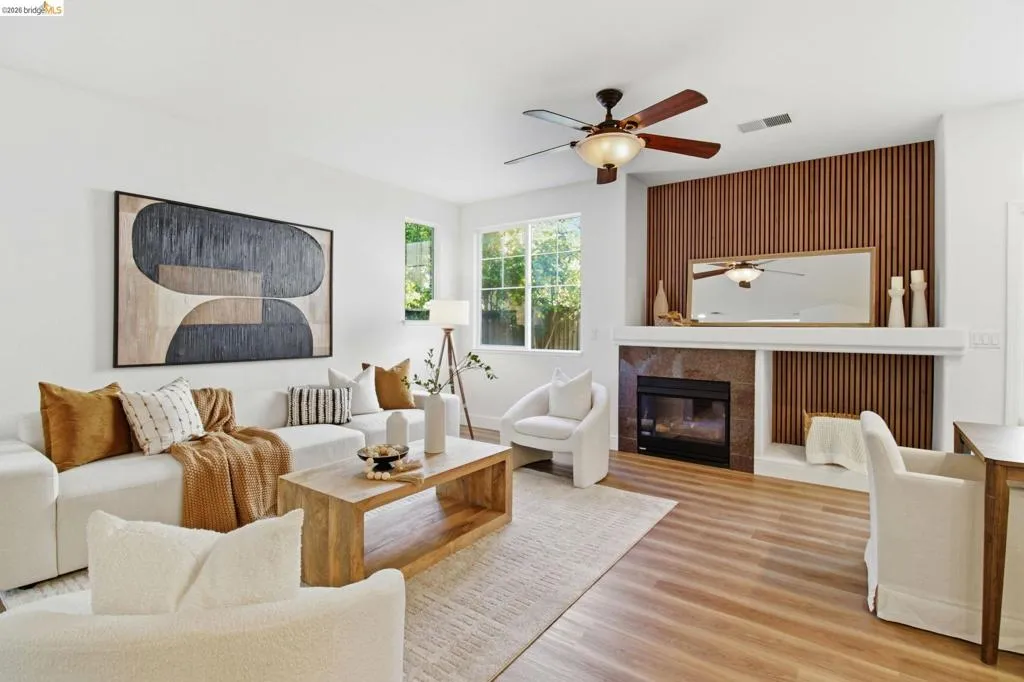 Bright living area featuring wood-finish flooring, a contemporary fireplace with a stone surround and wood slat accent wall, and a ceiling fan