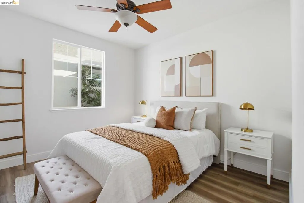 Bright interior space featuring a window with exterior views, wood-finish flooring, a ceiling fan with wood-tone blades, and white walls