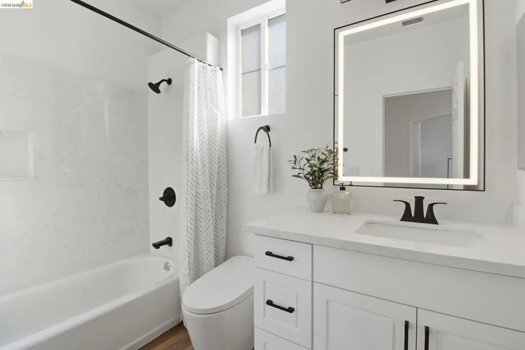 Bright bathroom featuring a white vanity with integrated sink, a rectangular illuminated mirror, matte black fixtures, and a white bathtub with a tile-look surround