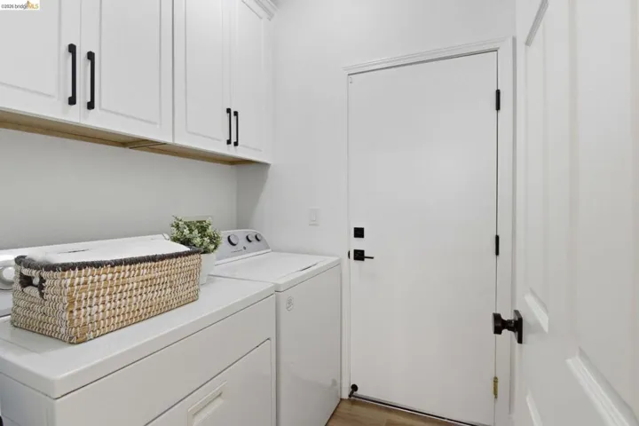Dedicated laundry area featuring white shaker-style cabinetry with matte black hardware, a wood-finish shelf, and light wood-finish flooring