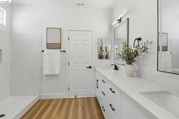 White bathroom featuring a dual vanity with undermount sinks, wood-finish flooring, and a walk-in shower with a low-profile base