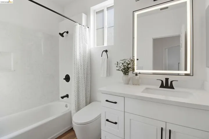 Bright bathroom featuring a white vanity with integrated sink, a rectangular illuminated mirror, matte black fixtures, and a white bathtub with a tile-look surround