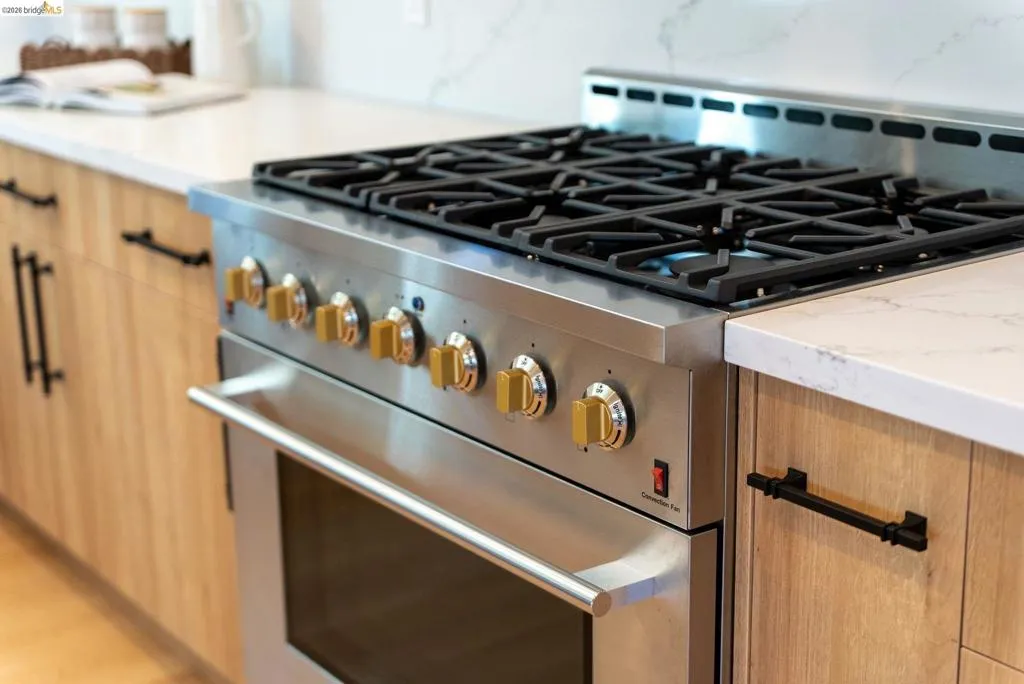 Stainless steel range with gold-toned knobs and cast iron grates, integrated into light wood-finish cabinetry with matte black hardware and white stone countertops