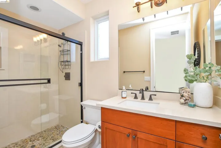 Bathroom featuring a glass-enclosed shower with pebble flooring, a single vanity with a light-colored countertop, dark wood-finish cabinetry, and a framed mirror with overhead lighting