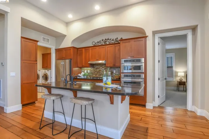 Gourmet kitchen featuring a large central island with seating, stainless steel appliances, rich wood cabinetry, stone tile backsplash, and wood-finish flooring