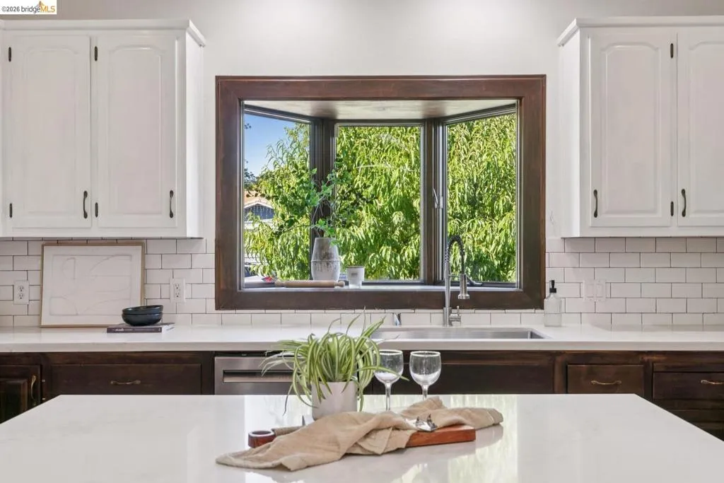 Kitchen featuring a bay window, white shaker-style upper cabinetry, dark wood-finish lower cabinetry, subway tile backsplash, and light-toned countertops