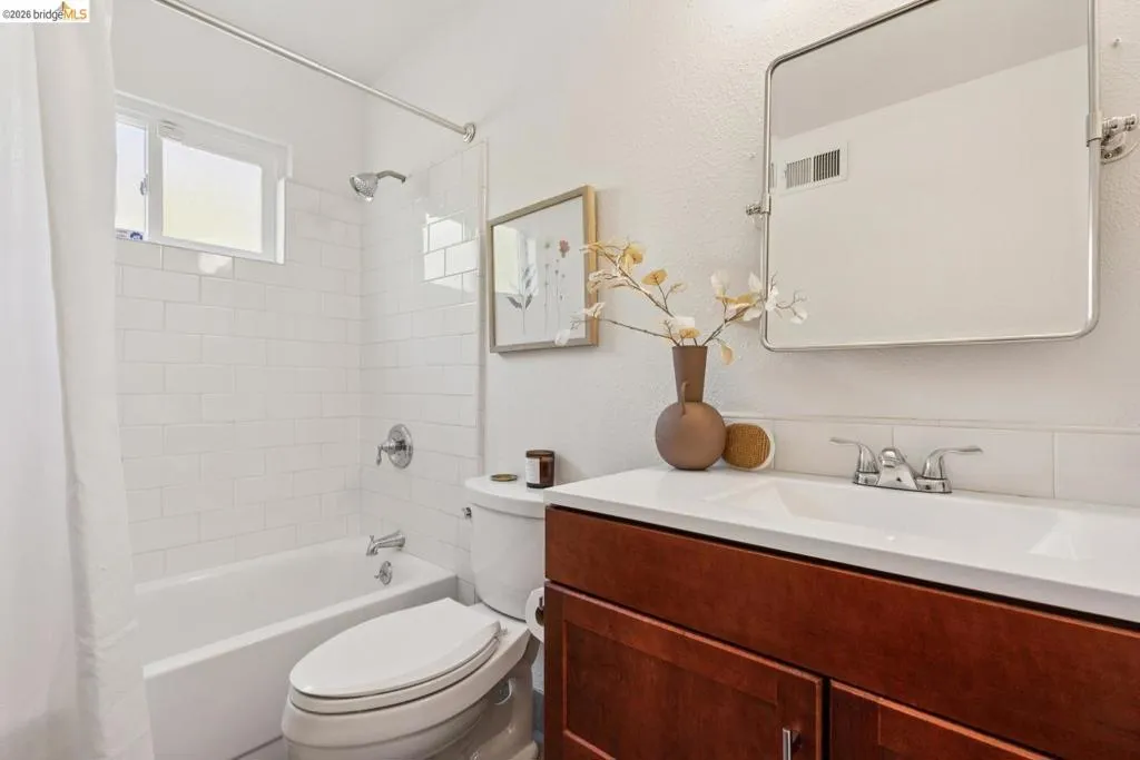 Bathroom featuring a white subway tile shower surround, a built-in tub, a single window, a wood-finish vanity with a white countertop and integrated sink, and a framed mirror