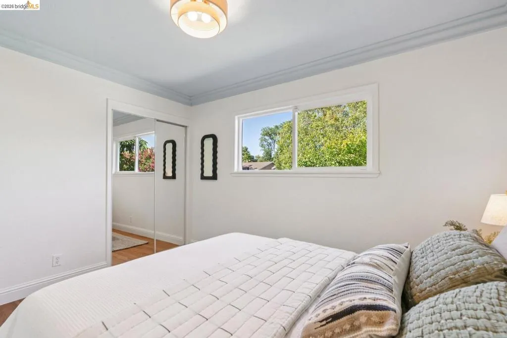 Bright bedroom featuring white walls, wood-finish flooring, a contemporary ceiling light fixture, and a window with green tree foliage outside