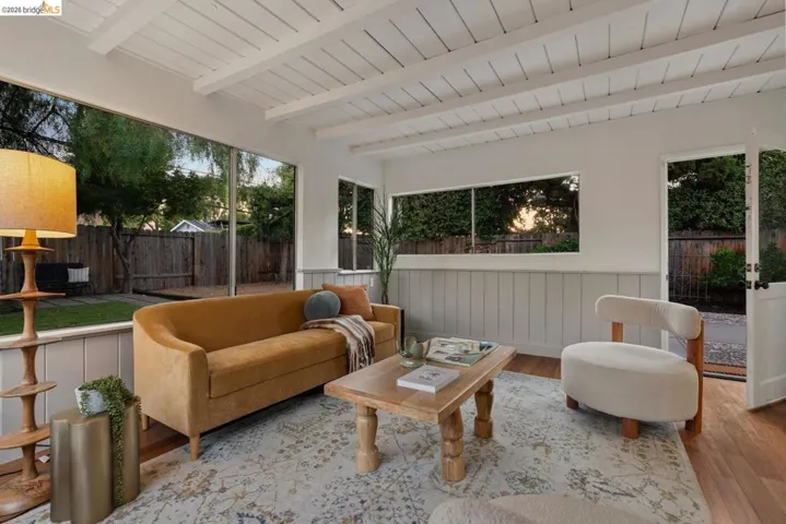 Living space with exposed white beam ceilings, expansive window overlooking the backyard, wood-finish flooring, and wainscoting