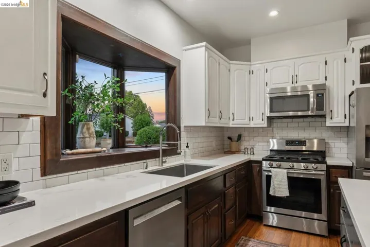 Kitchen featuring a wood-framed bay window, white shaker-style upper cabinetry, dark wood lower cabinetry, white subway tile backsplash, and stainless steel appliances including a gas range and microwave