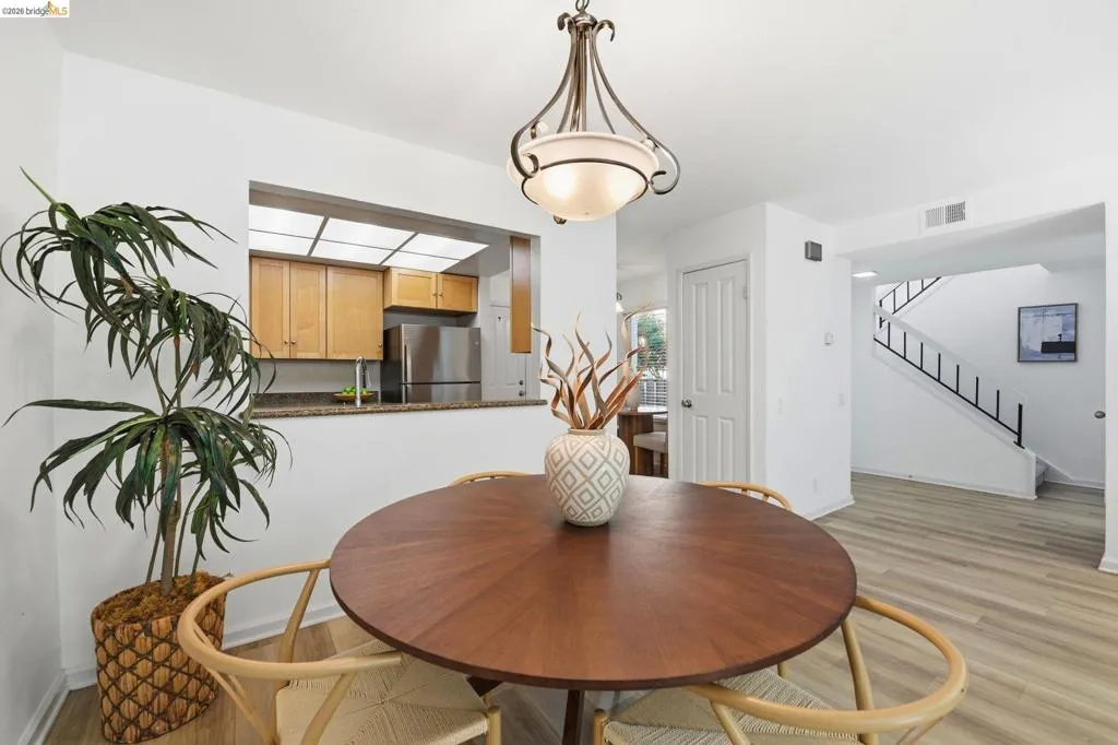 Dining area featuring a contemporary chandelier, wood-finish flooring, and a kitchen pass-through with light wood cabinetry, stone-look countertops, and stainless steel appliances