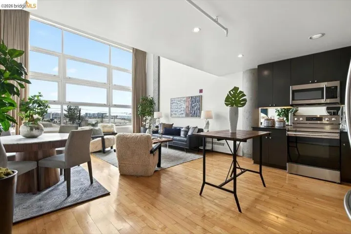 Expansive open-concept living area featuring a full-height window wall with multiple panes, light wood-finish flooring, and a kitchen area with dark cabinetry, stainless steel appliances, and a concrete structural pillar