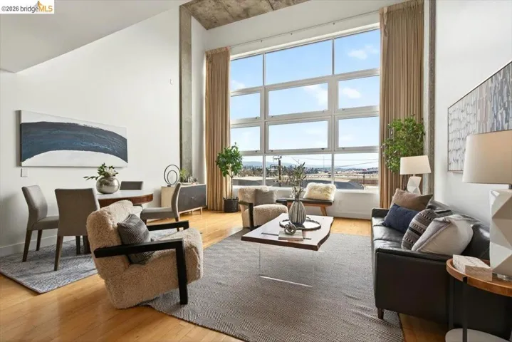 Spacious open-concept living area featuring a large grid-pane window, high ceilings with exposed concrete, wood-finish flooring, and a dining area