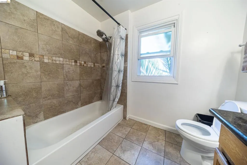 Bathroom featuring a tiled shower surround with decorative accent band, a white tub, a white framed window, and tiled flooring