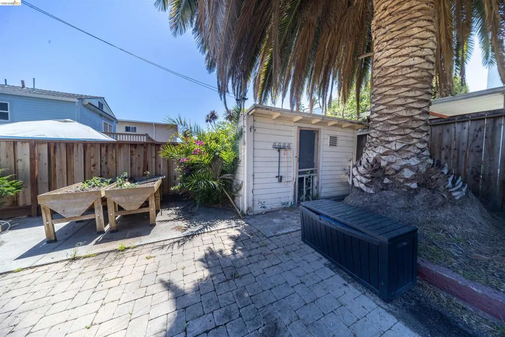 Paved patio area featuring brick-patterned hardscaping, a mature palm tree, a wooden privacy fence, and a white utility structure