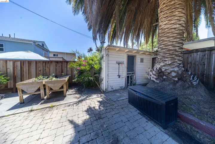 Paved patio area featuring brick-patterned hardscaping, a mature palm tree, a wooden privacy fence, and a white utility structure