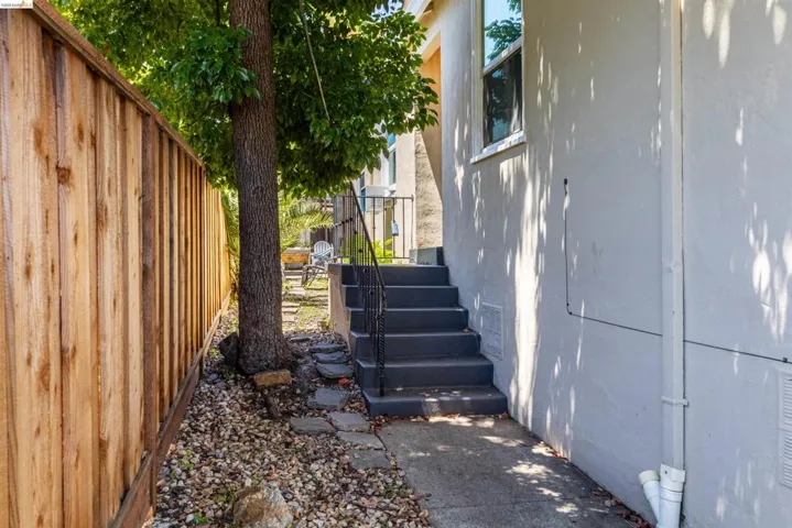 Exterior walkway featuring concrete steps with a black metal handrail, a light-colored stucco wall, a wood plank fence, and a mature tree providing natural shade