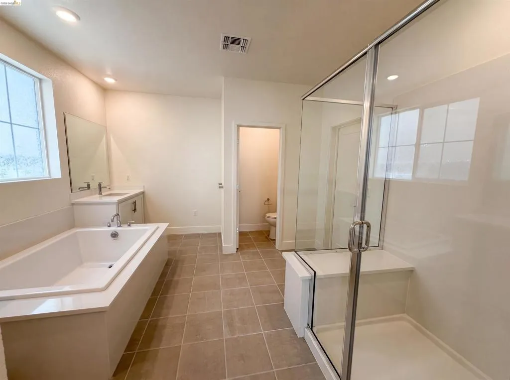 Spacious bathroom featuring a built-in soaking tub with a surrounding deck, a separate glass-enclosed shower with a bench, a single vanity with a solid surface countertop, and large format tile flooring