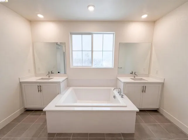 Recessed lighting illuminates a built-in soaking tub with chrome fixtures, flanked by two white vanity cabinets featuring undermount sinks, chrome faucets, and quartz countertops