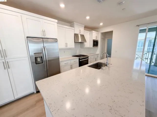 Kitchen featuring a large central island with an undermount sink and chrome faucet, white shaker-style cabinetry, stainless steel appliances, and wood-finish flooring