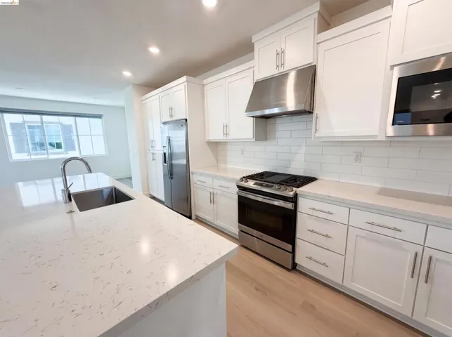 Modern kitchen featuring a large island with an undermount sink, stainless steel appliances, white shaker cabinetry, white subway tile backsplash, and light wood-finish flooring