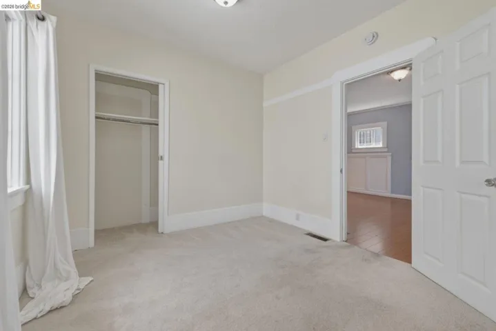 Neutral-toned room with plush carpeting, featuring a window with white drapes, a built-in closet, and a doorway opening to an additional room with wood-finish flooring