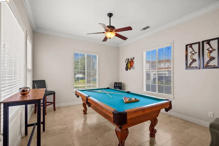 Spacious room featuring neutral wall tones, crown molding, two large windows with blinds, a ceiling fan with integrated lighting, and tile flooring
