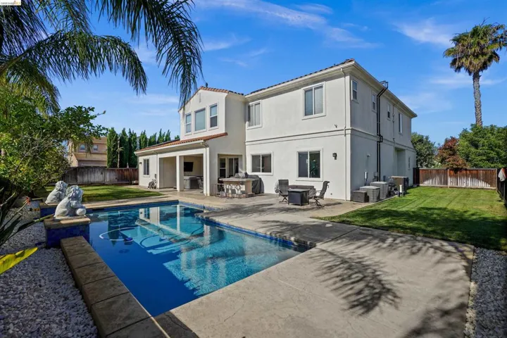 Rear exterior featuring a rectangular in-ground pool with concrete surround, covered patio with built-in outdoor kitchen, stucco siding, and a fenced yard with mature landscaping