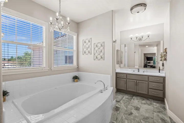 Bathroom featuring a built-in soaking tub with white tile surround, dual windows with blinds, a crystal chandelier, a vanity with dual sinks and a large mirror, and gray-toned floor tiling