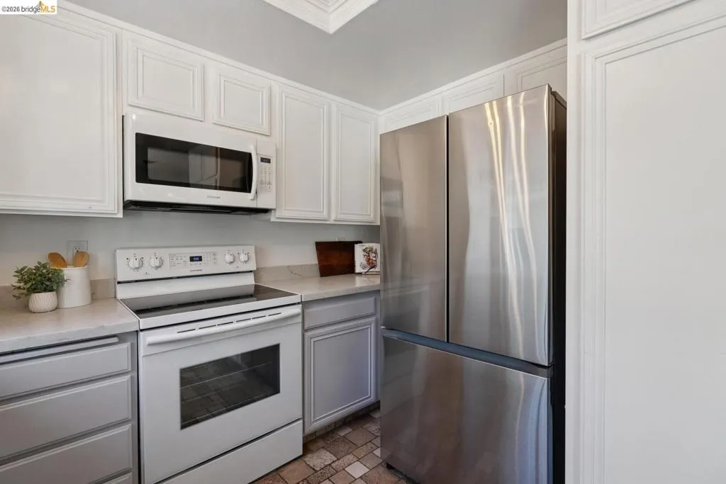 Kitchen featuring white upper cabinetry with crown molding, light blue lower cabinetry, a stainless steel refrigerator, white electric range, and a built-in white microwave