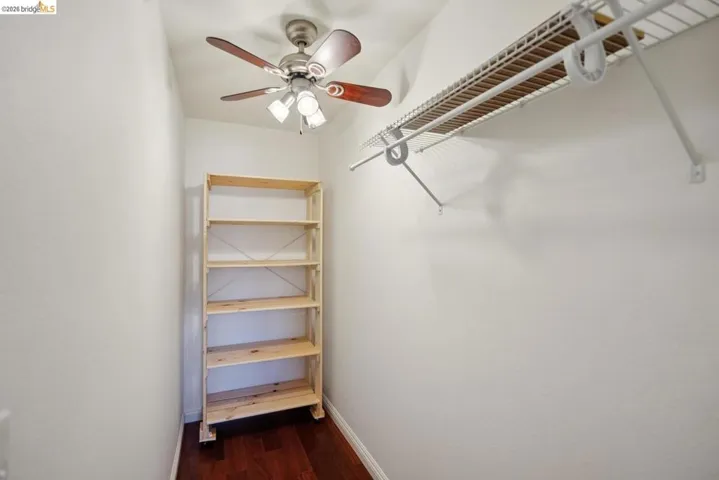 Compact room featuring a ceiling fan with integrated lighting, wood-finish flooring, a freestanding six-shelf unit, and a wall-mounted wire shelving system