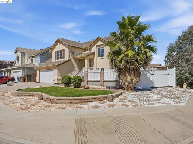 Stucco exterior with a two-car garage, mature palm tree, decorative stone landscaping, and a white privacy fence