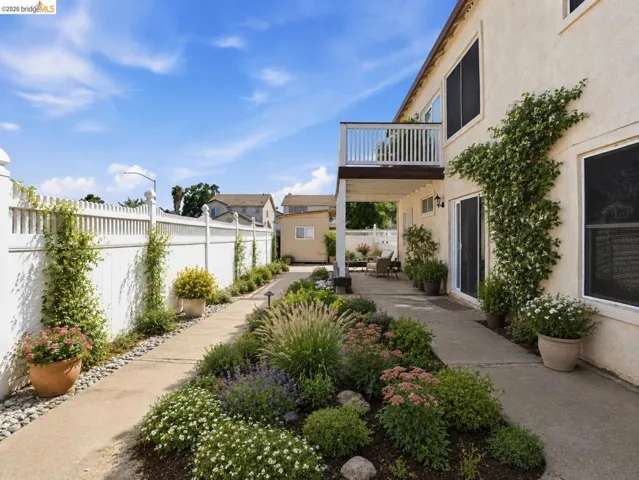 Exterior patio area featuring a second-story balcony with white railing, a concrete walkway, and extensive landscaping with various plants