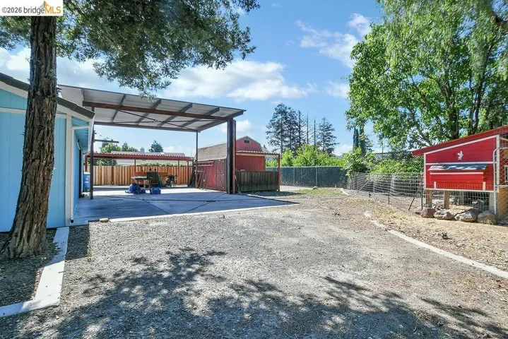 Expansive gravel lot featuring a large metal-framed carport with a concrete slab, a light blue storage structure, a red outbuilding, and mature trees