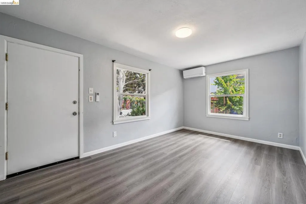 Interior room featuring light gray walls, wood-finish flooring, two windows with white trim, and a wall-mounted mini-split HVAC unit