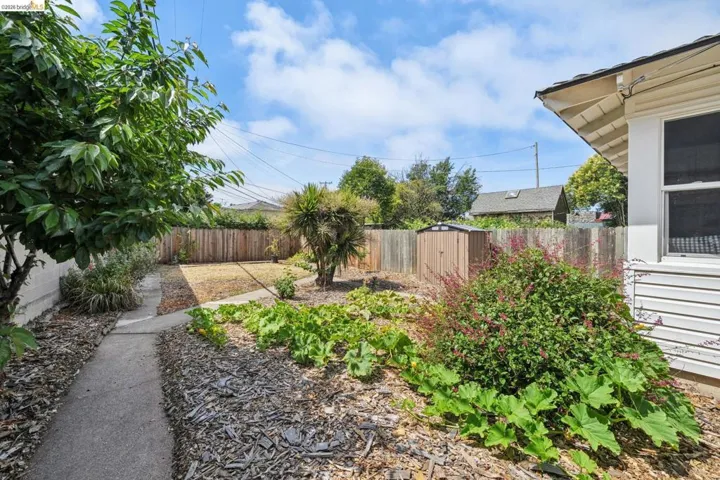 Fenced backyard featuring a concrete pathway, a storage shed, established trees, and planting beds