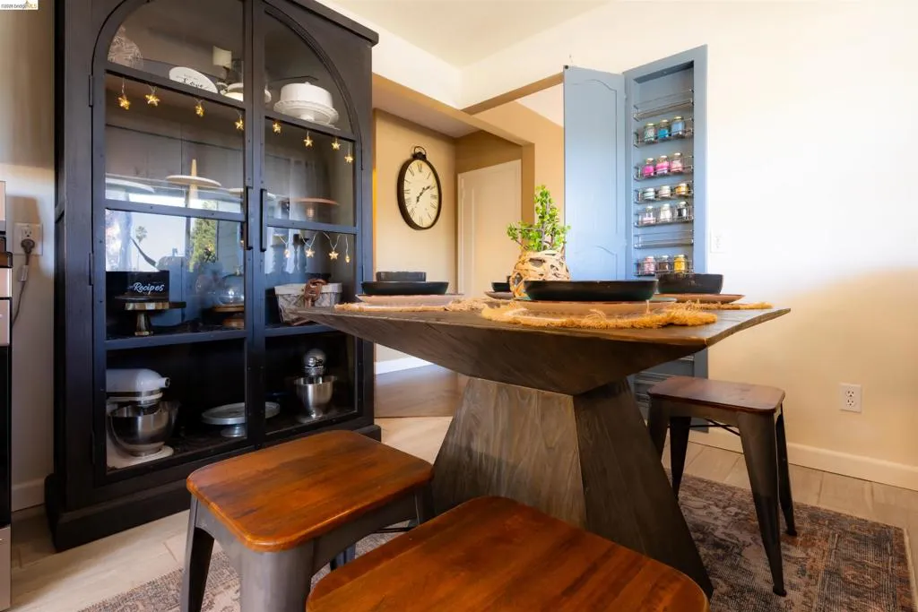 Dining area featuring a modern wood-finish table with a unique pedestal base, light-toned flooring, and a built-in ironing board nook, repurposed as a sprinkle/spice rack